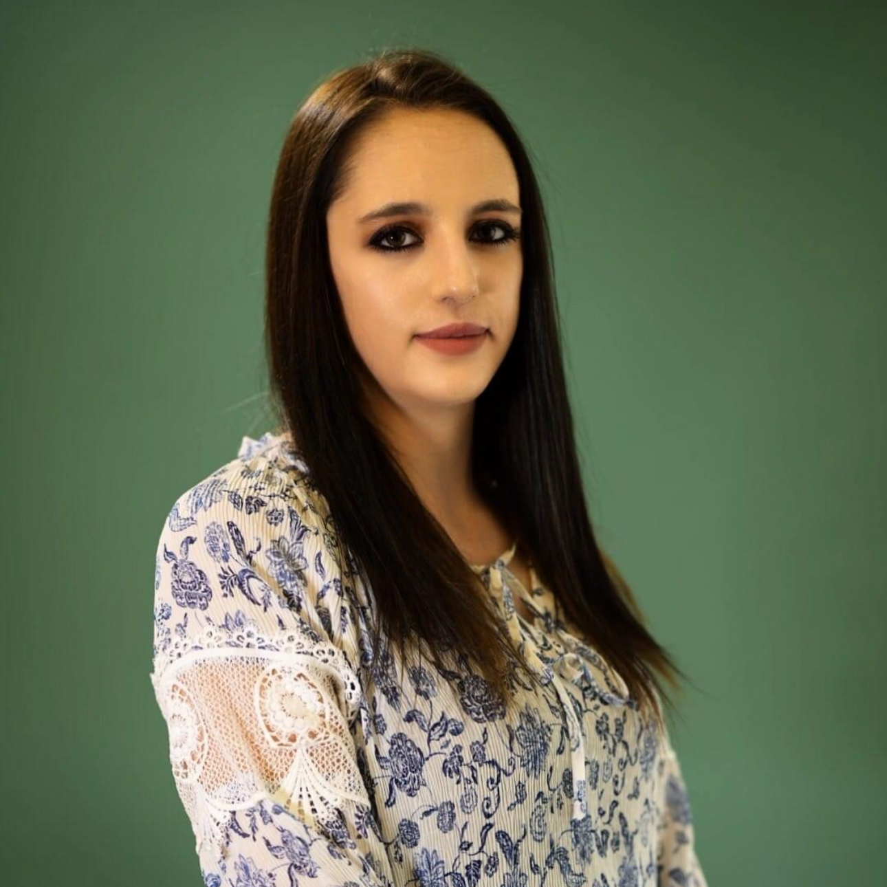 A woman in a floral shirt is standing in front of a green background.