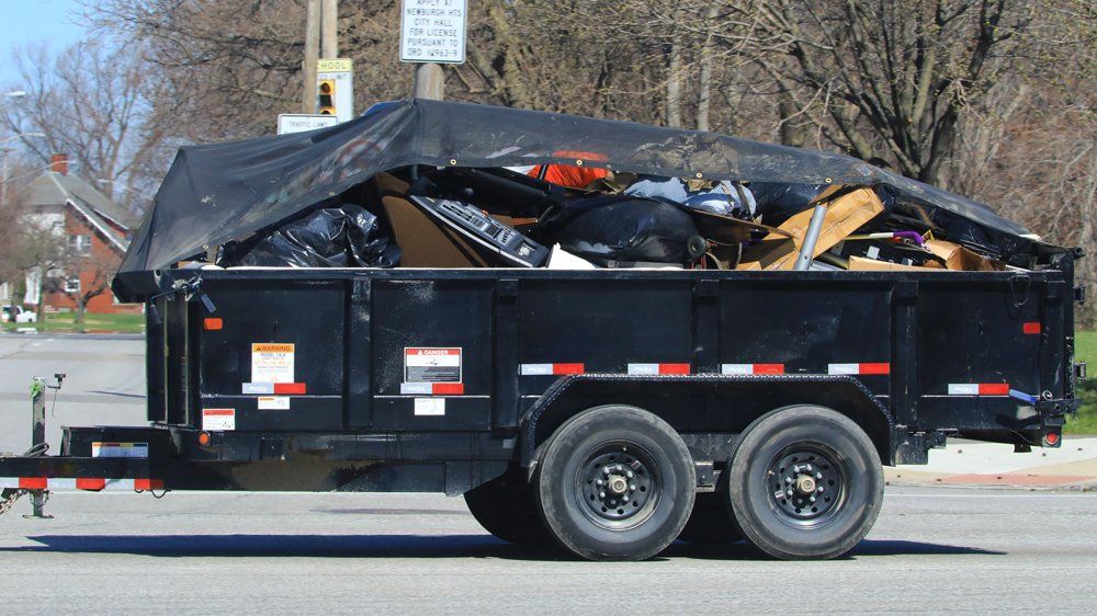 A black trailer filled with assorted junk and debris, covered with a tarp, parked on a street. This image represents the kind of load handled by Abate Rubbish Removal's junk removal services in Newcastle.