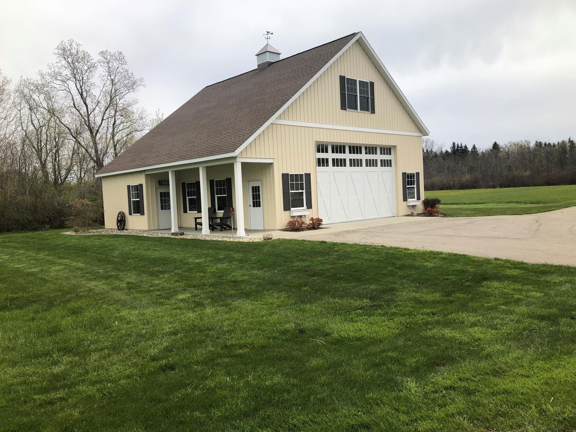 Tan two-story barn with white garage door, columns, dark shutters, and a brown roof, set on green grass.