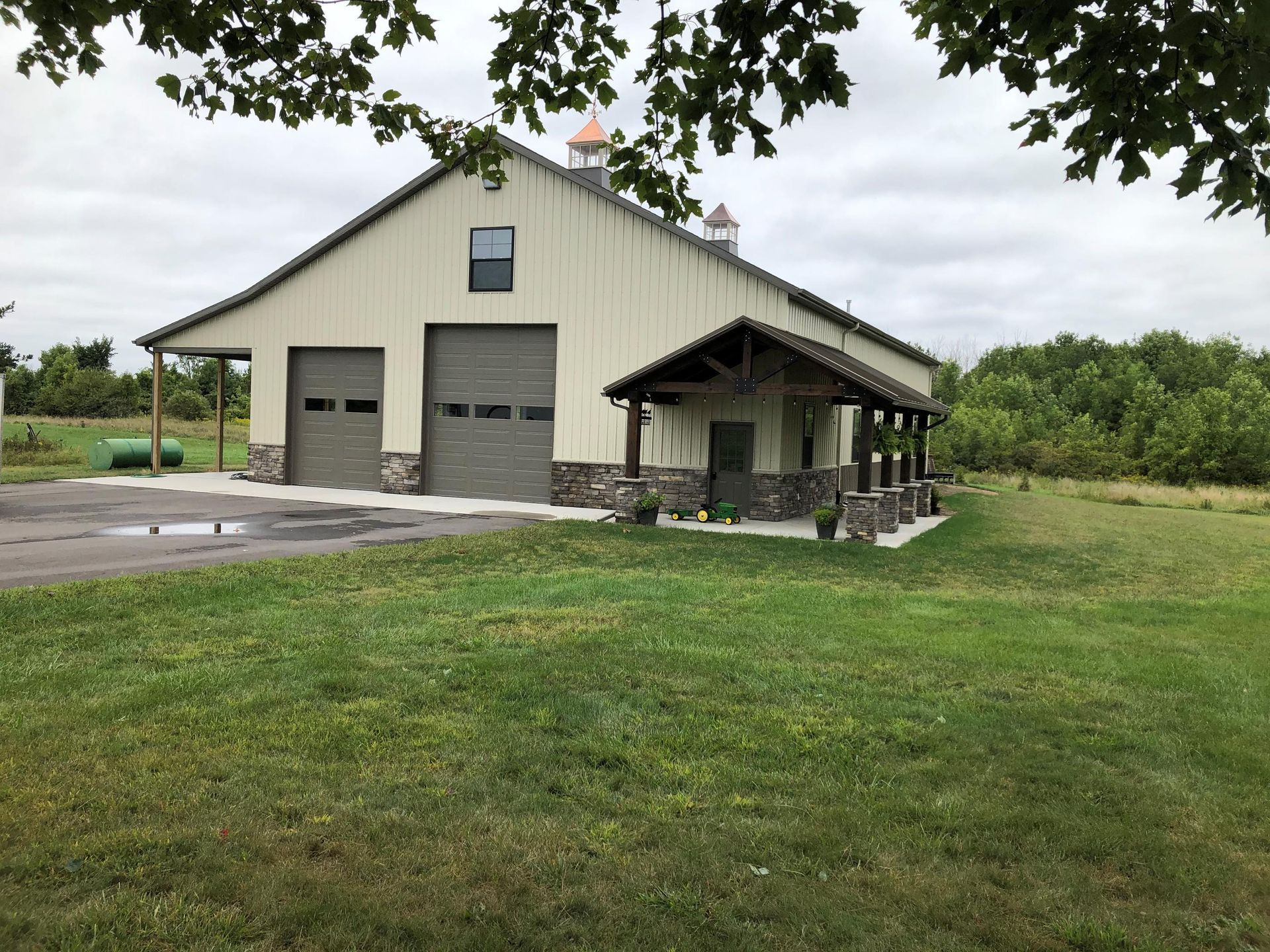 Tan barn with stone accents, two garage doors, and a covered porch on a grassy field under an overcast sky.