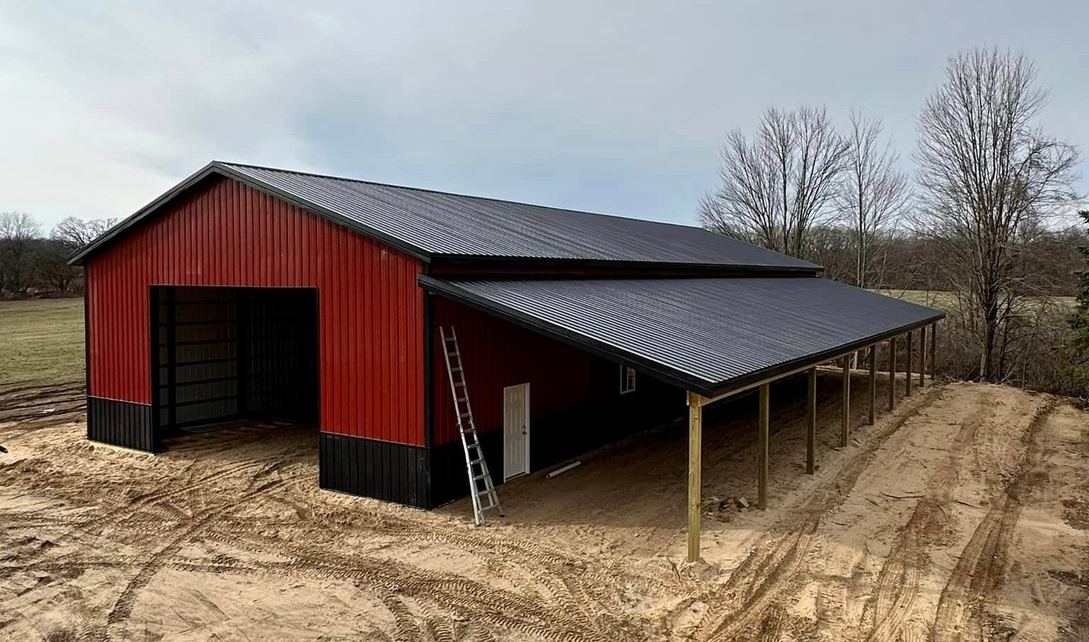 Red and black metal barn with a dark roof and a lean-to roof extension. A ladder leans against the building.