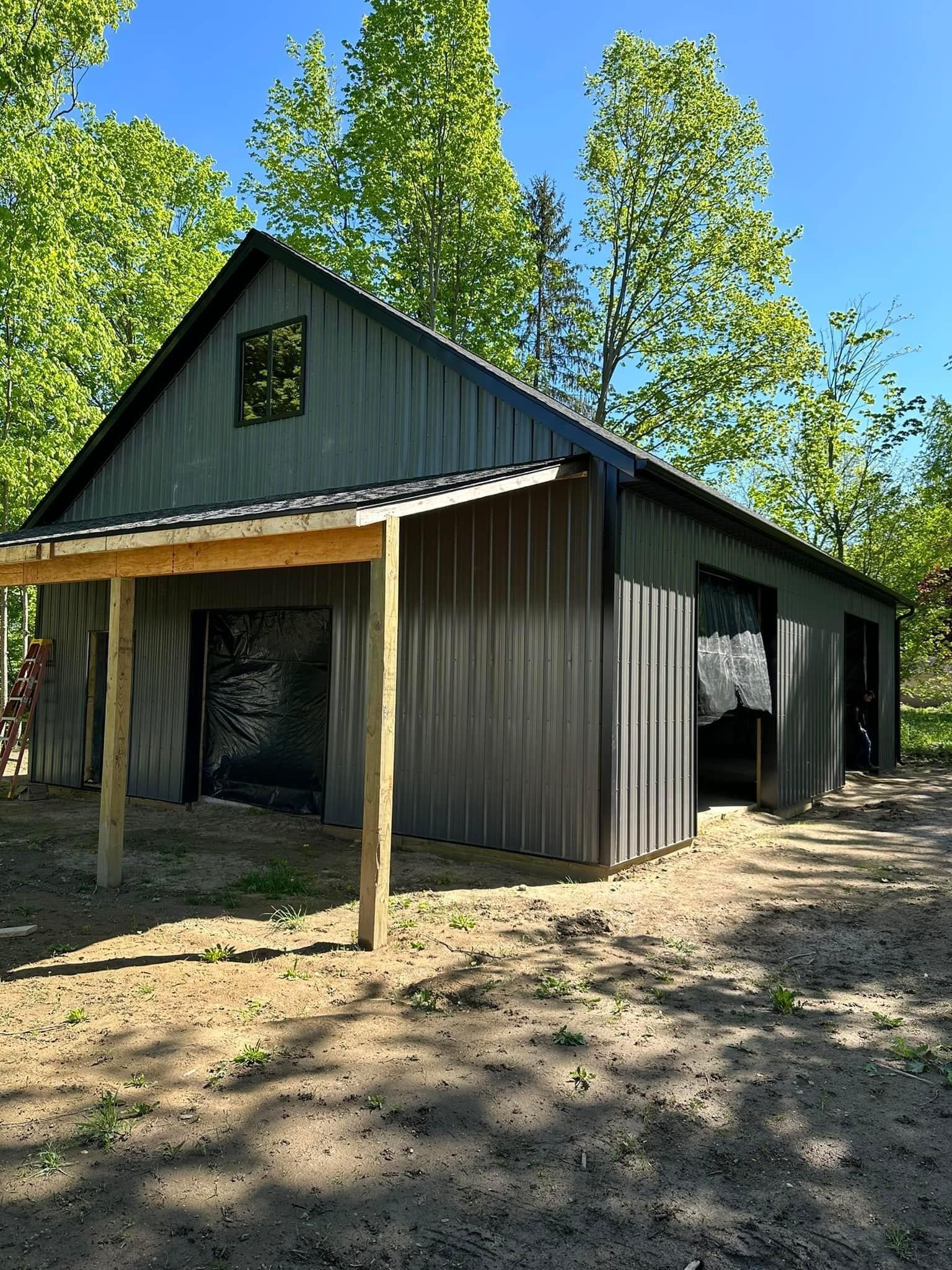 Gray metal barn with a small porch, built on a dirt lot, surrounded by trees.