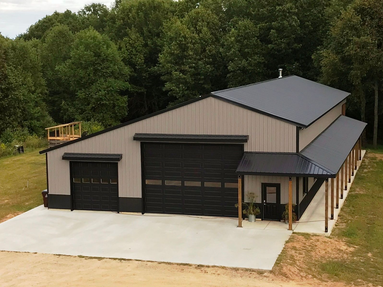 Gray and black metal building with two garage doors and a porch, set in front of trees.