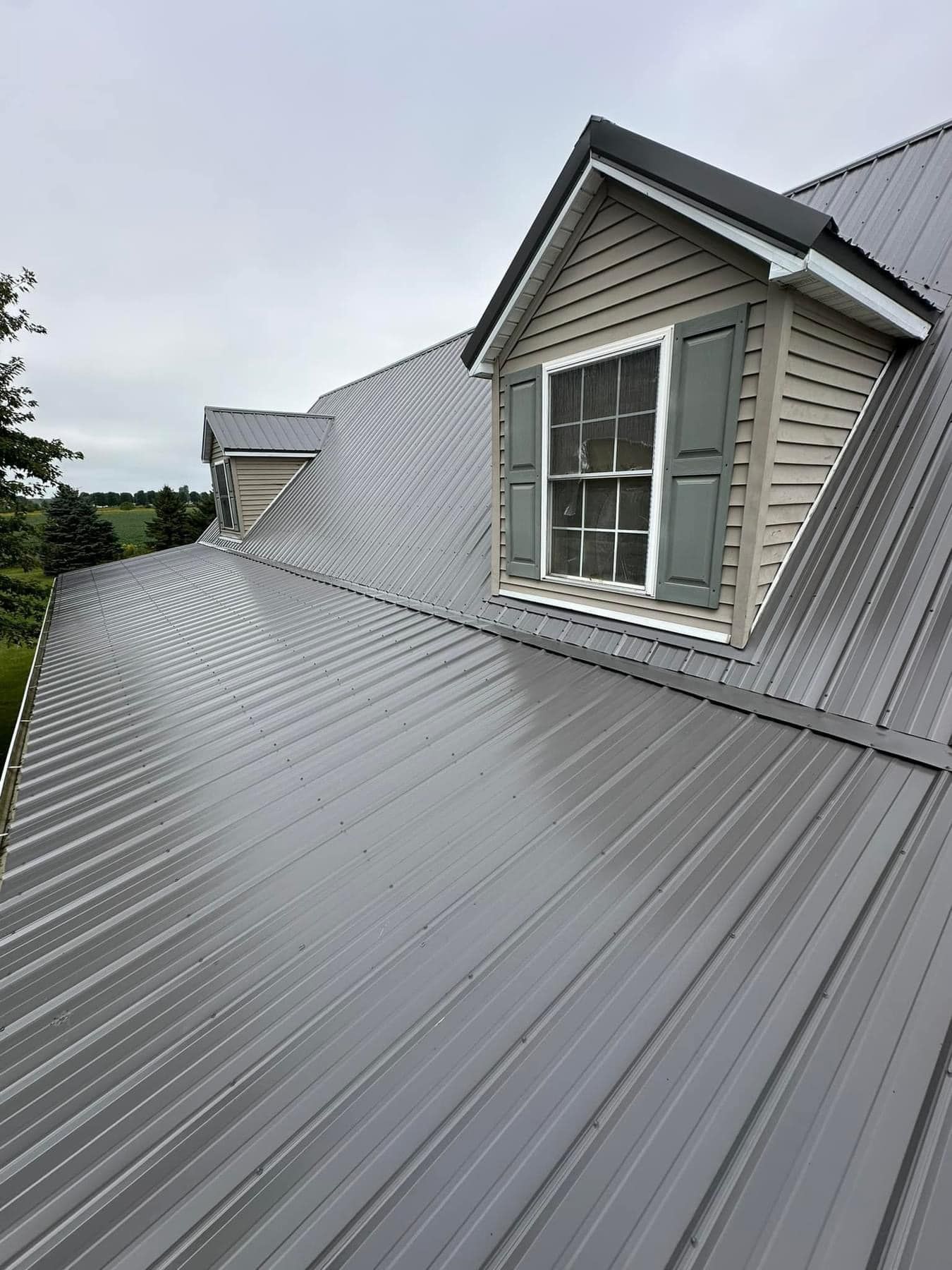 Gray metal roof with dormer windows, green shutters, on a cloudy day.