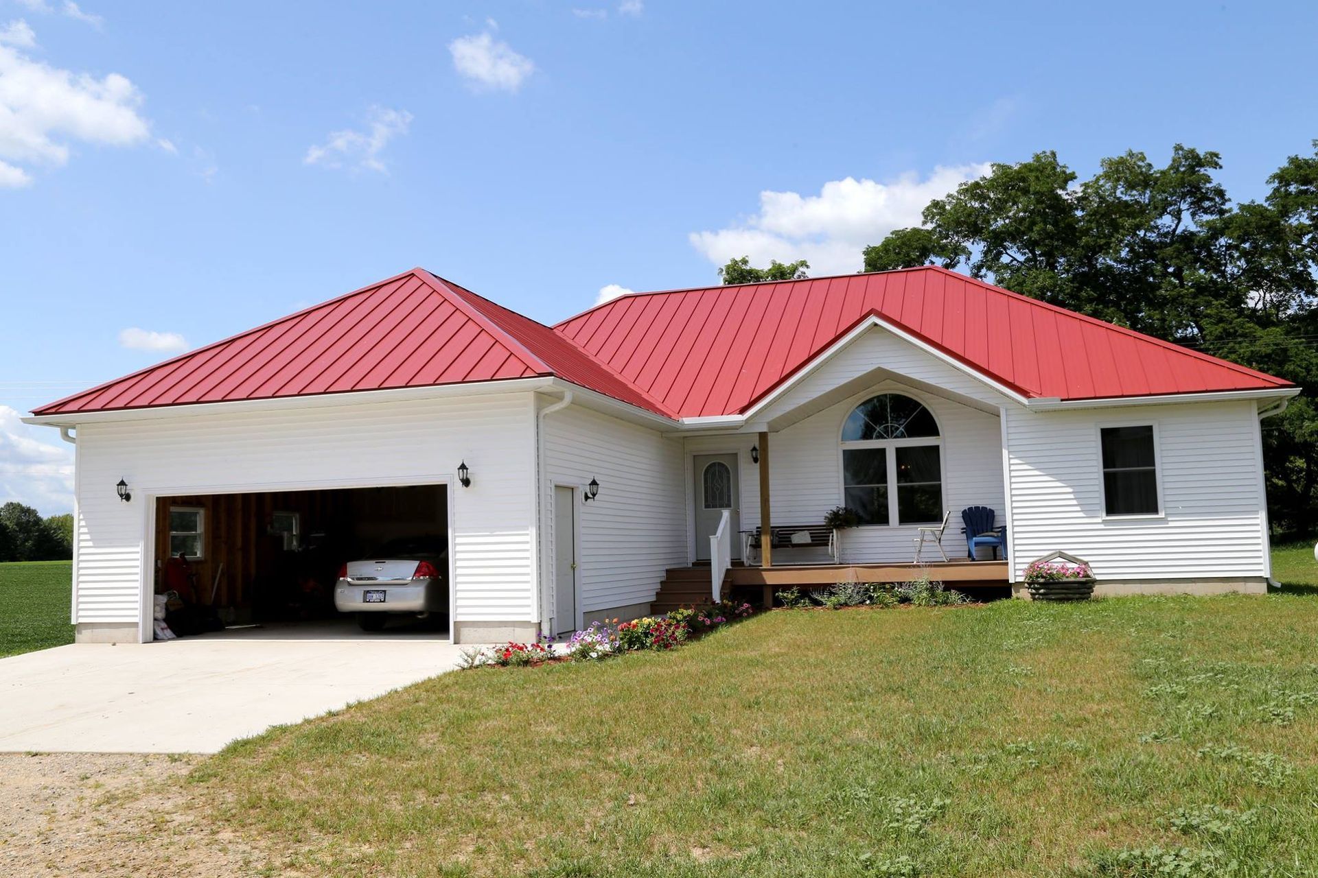 White house with red metal roof, attached garage, and small porch on a grassy hill under a blue sky.