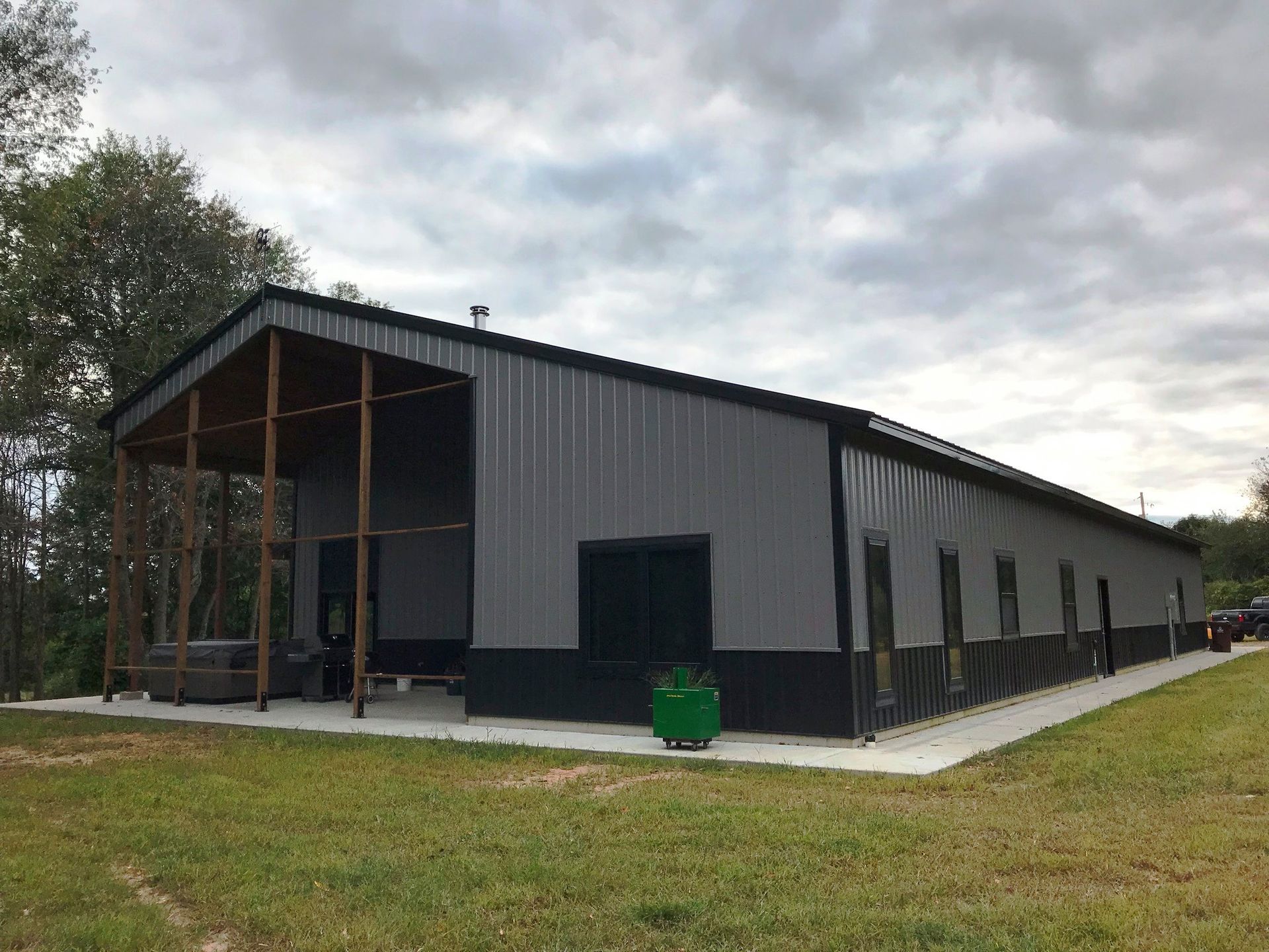 Gray metal building with black trim on a concrete slab, porch area, green trash can, overcast sky.