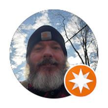 Man with a gray beard and black hat outside under a cloudy sky, with a white and orange star logo.
