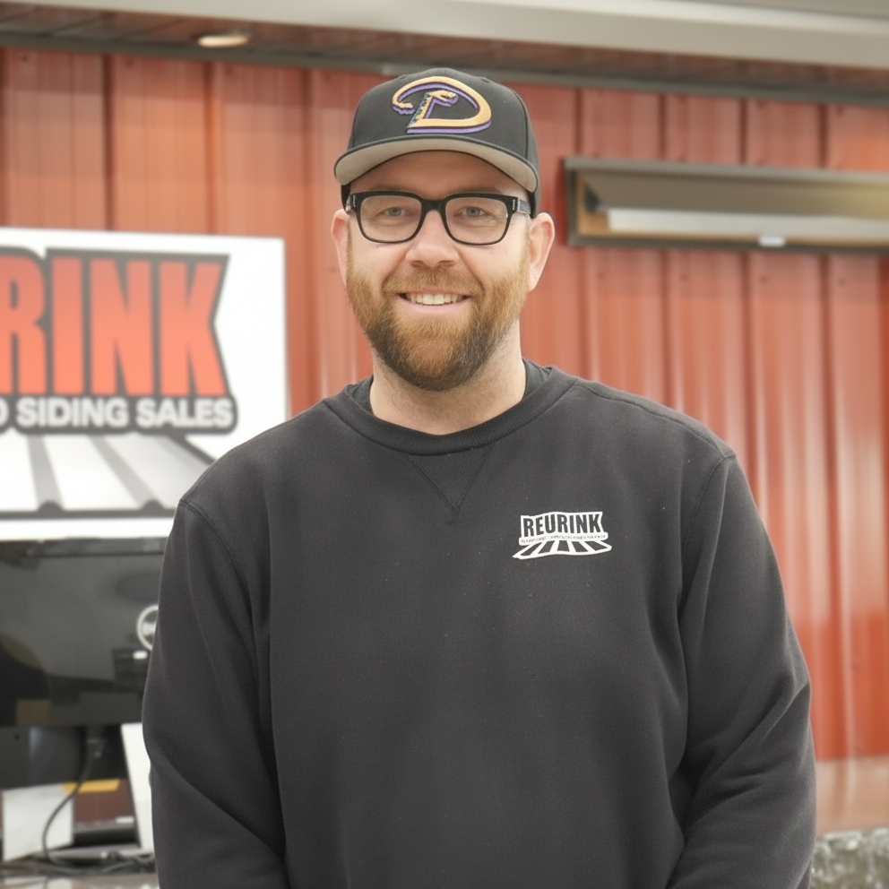 Man wearing glasses and a baseball cap smiling in front of a sign that says