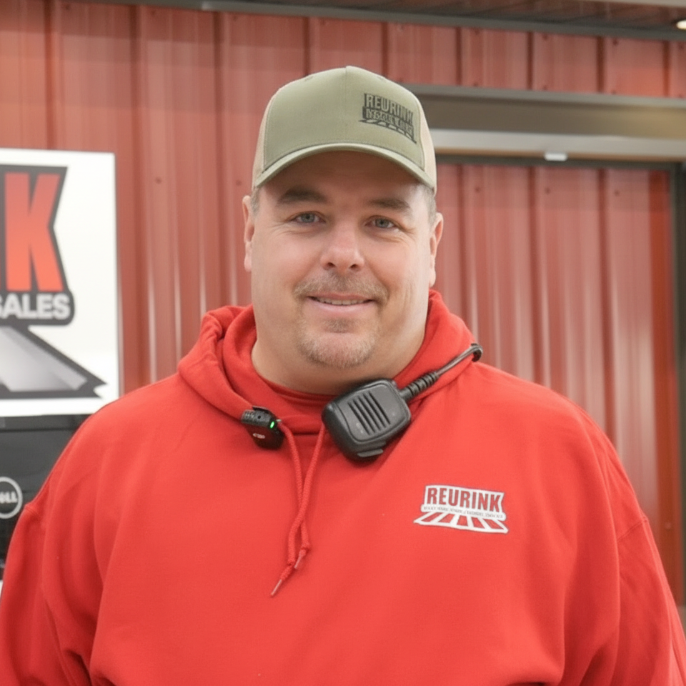 Man in red sweatshirt and green cap, smiling, with a radio mic. Standing in front of a building.
