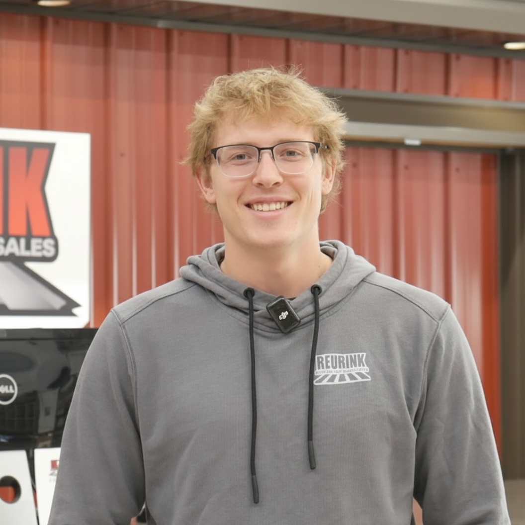 Young man with glasses and blonde hair wearing a gray hoodie, smiling in front of a red building.