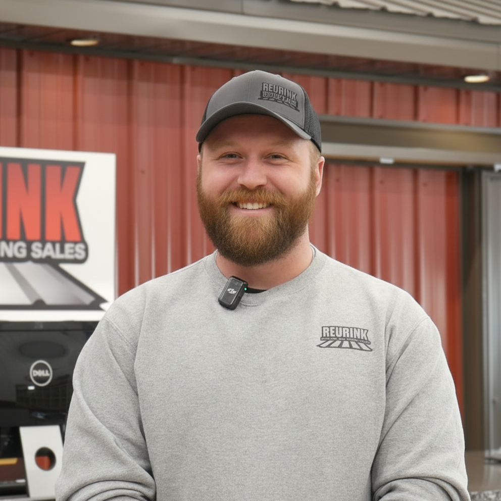 Man wearing a gray hat and sweatshirt smiles. He is standing outside a building with a sign that reads