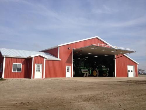 Red farm building with open bay door, housing a tractor; set on a gravel lot under a blue sky.