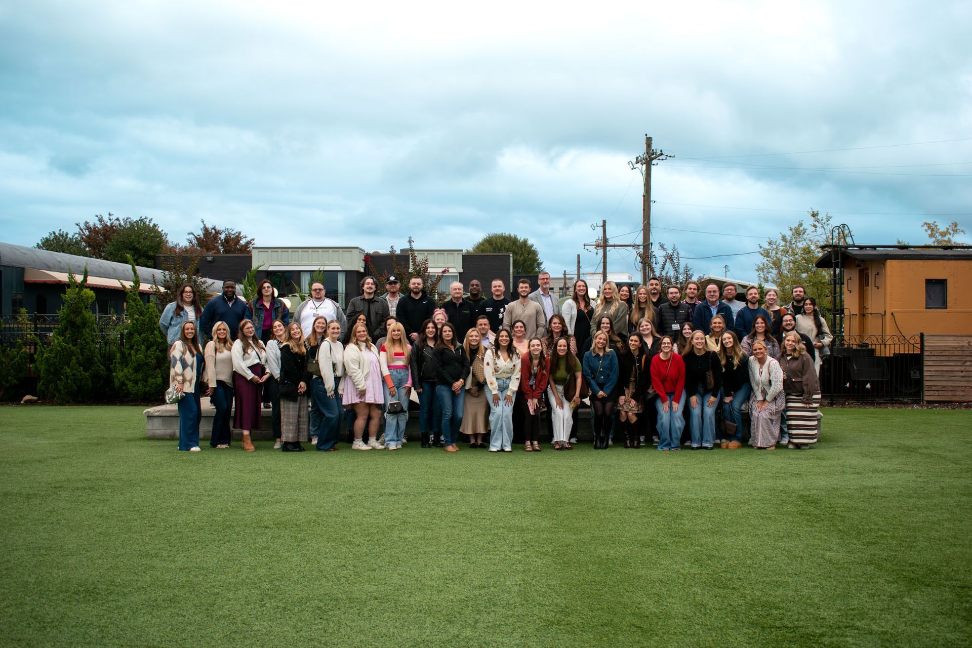 Large group of people standing on green grass, outdoors. Cloudy sky in the background, buildings and trees are visible.