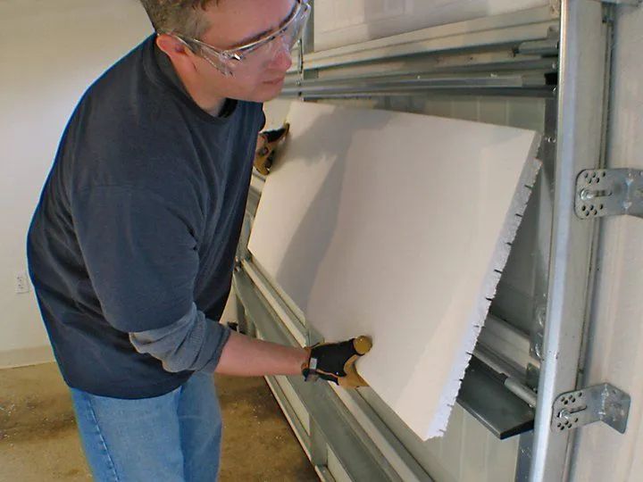 Man installing white insulation panel into a garage door. Wearing safety glasses and gloves.