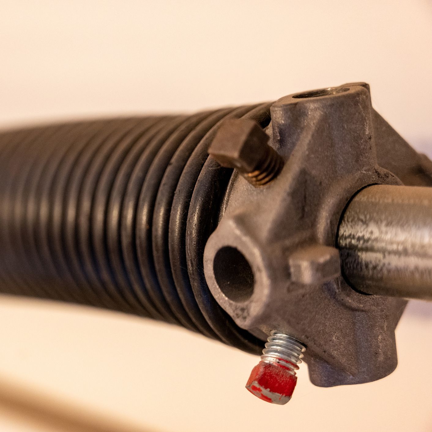 Close-up of a black garage door torsion spring coiled around a metal rod, with a metal bracket and red safety bolt.
