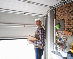 Man in hard hat inspects garage door, holding clipboard. Brick wall, open door, and white ceiling.