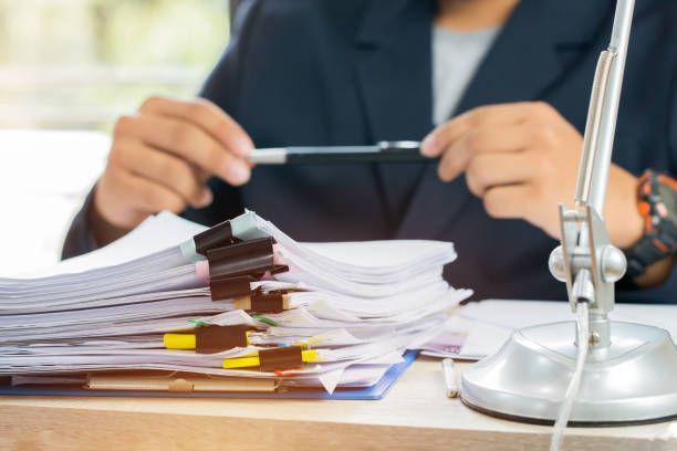 A person is sitting at a desk holding a pen over a pile of papers.