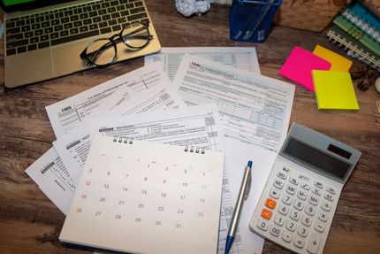 A desk with a laptop , calculator , calendar , pen , glasses and sticky notes.