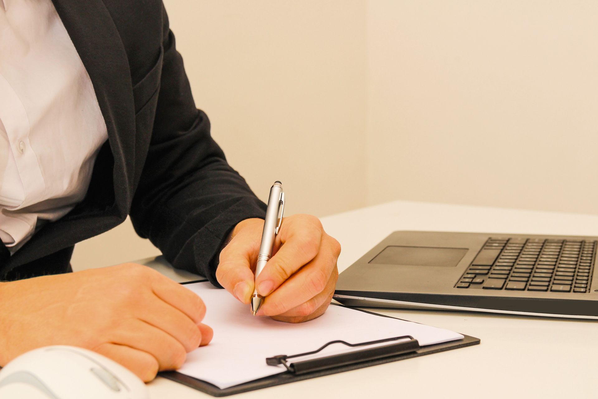 A woman is writing on a clipboard next to a laptop.