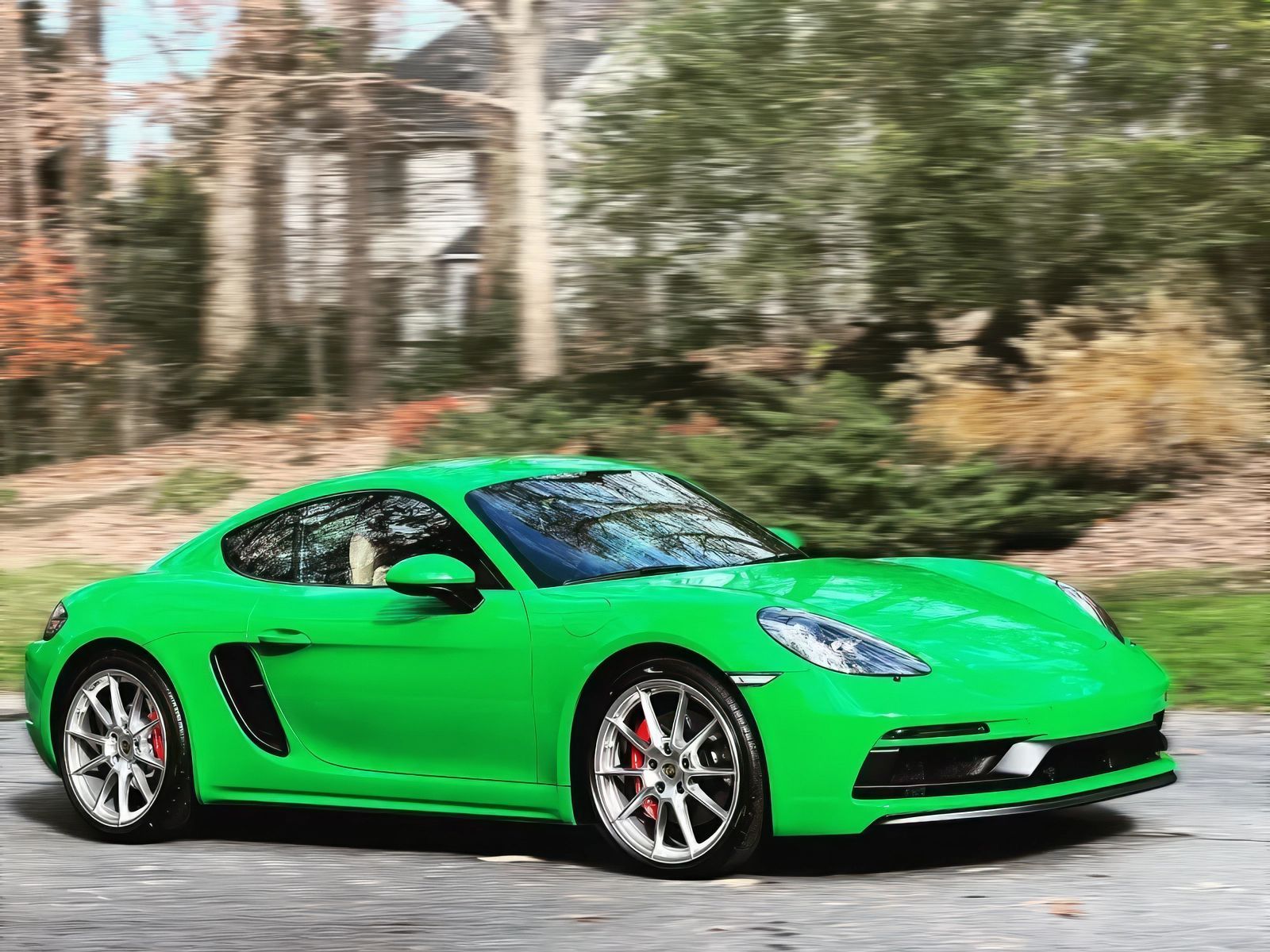 Bright green Porsche sports car driving on a road near trees and a house.
