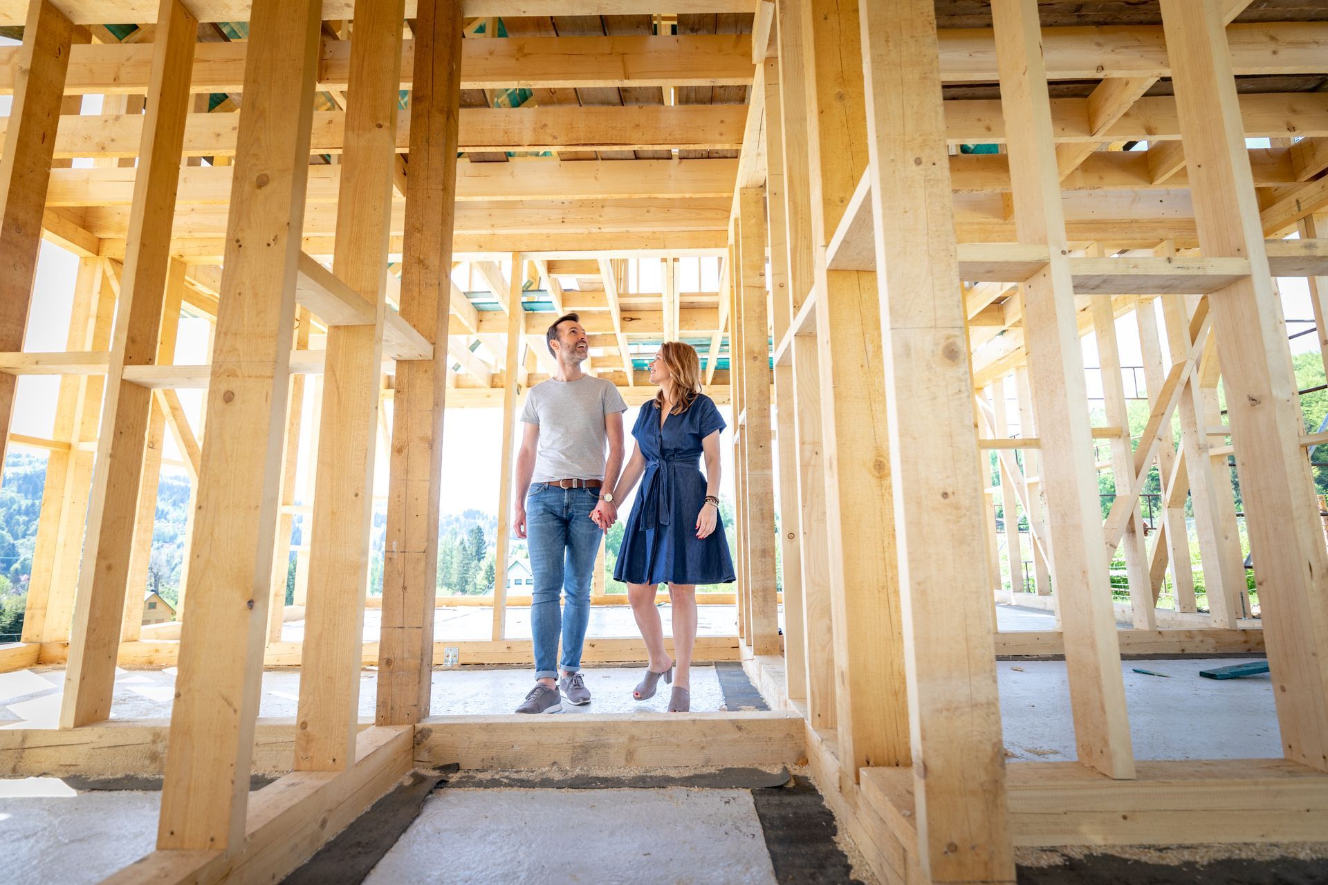 Two people stand in a wooden house frame under construction, looking at the camera.