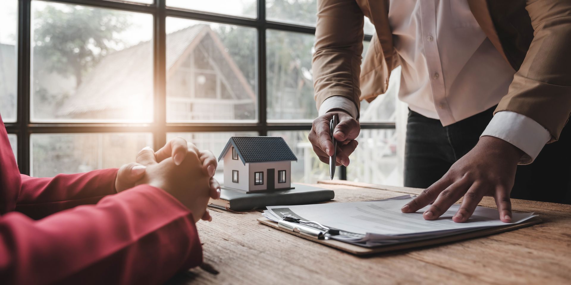 Person signing real estate documents beside a house model at a sunlit desk