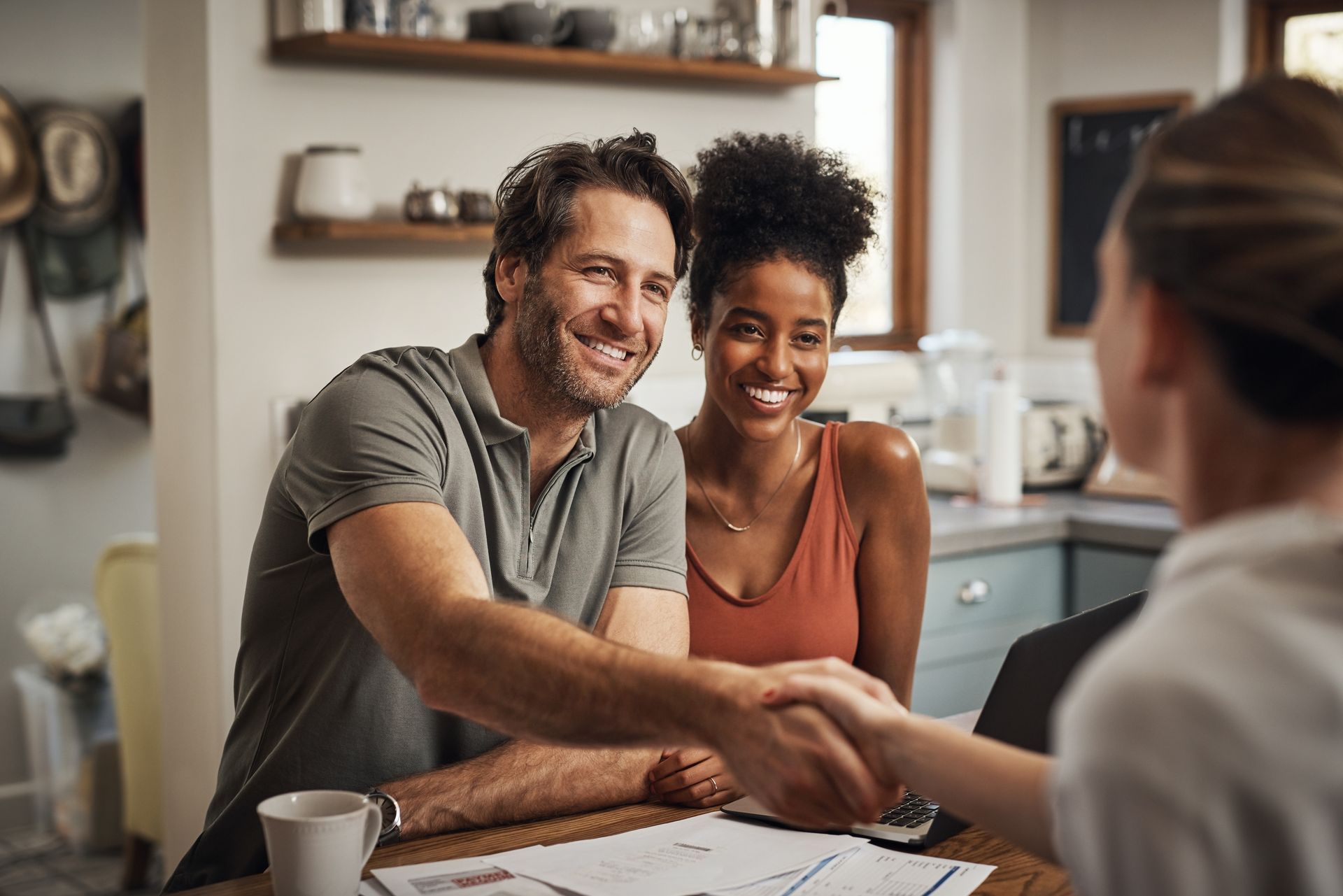 Two people shaking hands across a table in a bright kitchen, smiling during a meeting