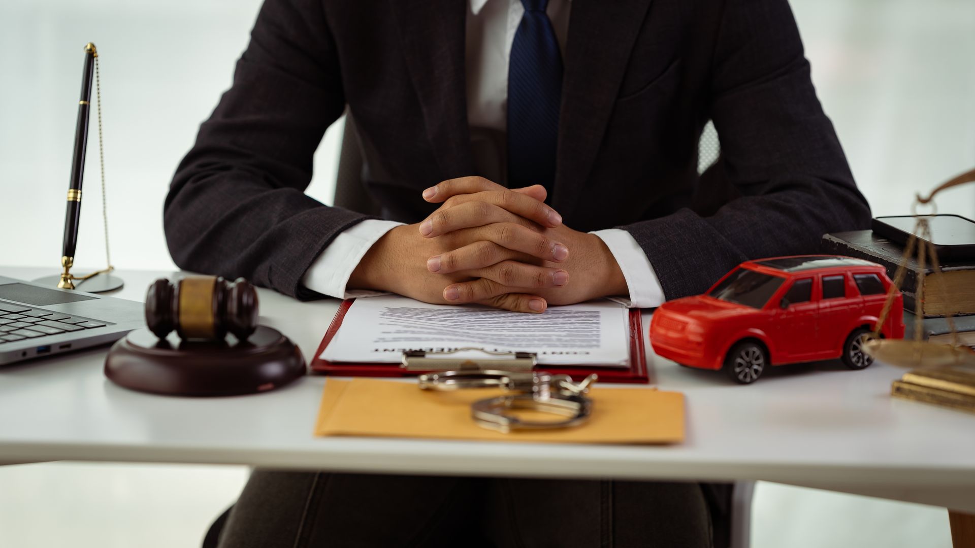 A person in a suit sits at a desk with a gavel, handcuffs, a red toy car, and legal documents.