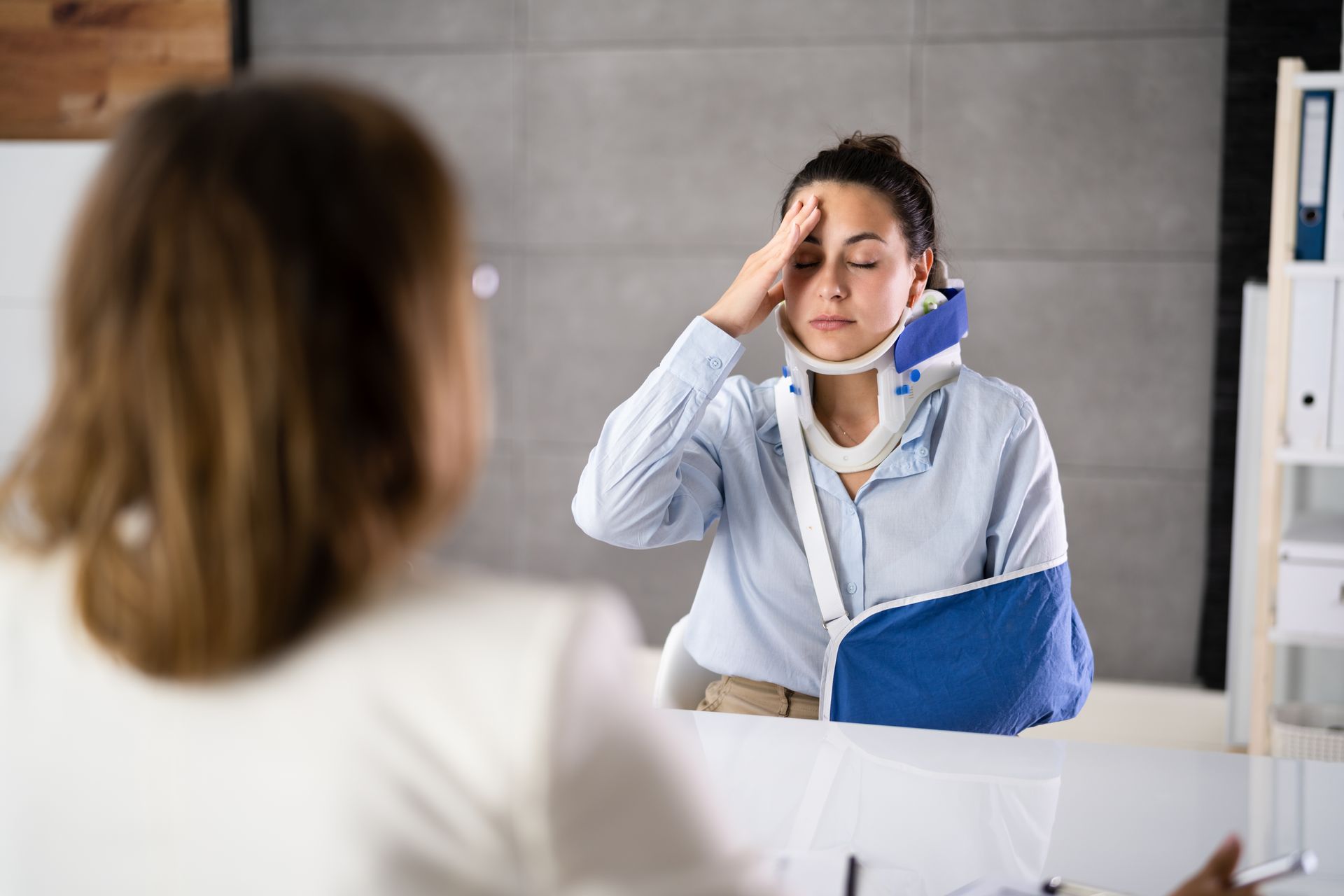 Woman with neck brace and arm sling, holding head, talking to someone at a desk.