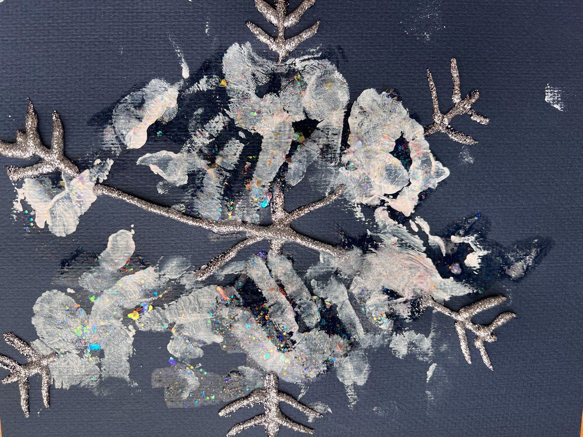 A six point child-made snowflake created with glitter glue and inward-pointing chevrons on the ends on black construction paper. The spines are decorated with handprints in white ink.