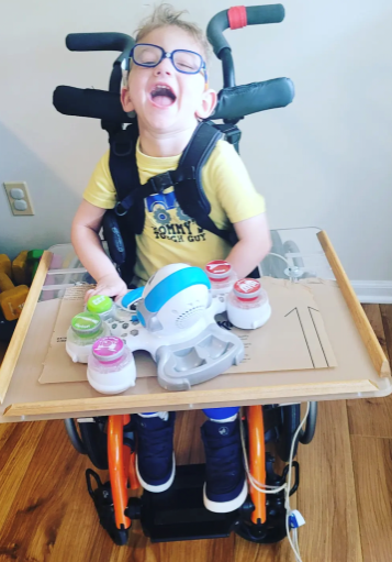 Child smiling at camera while sitting in wheelchair with tray and toy.