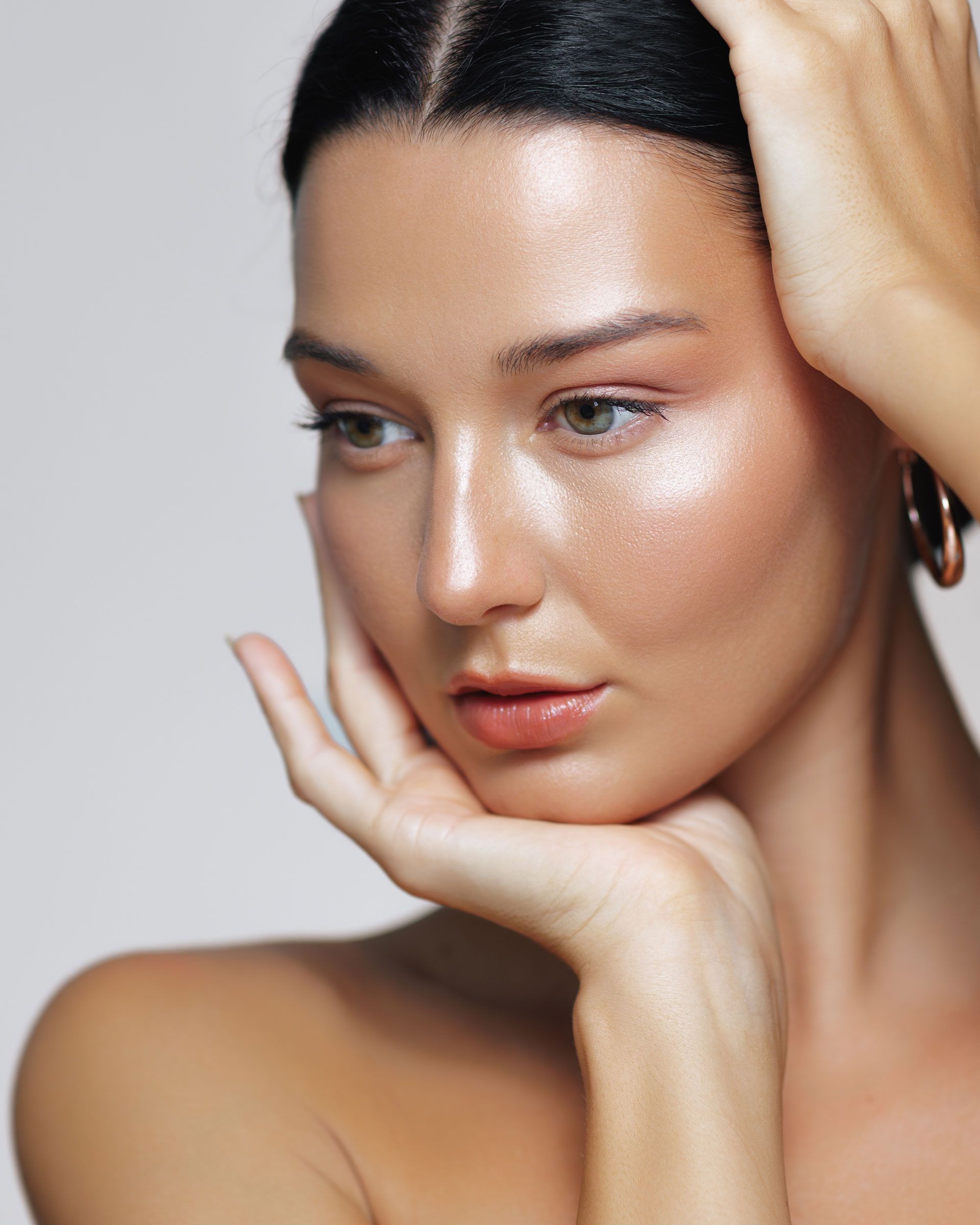 Woman with glowing skin, hand on face, looking down, light background.