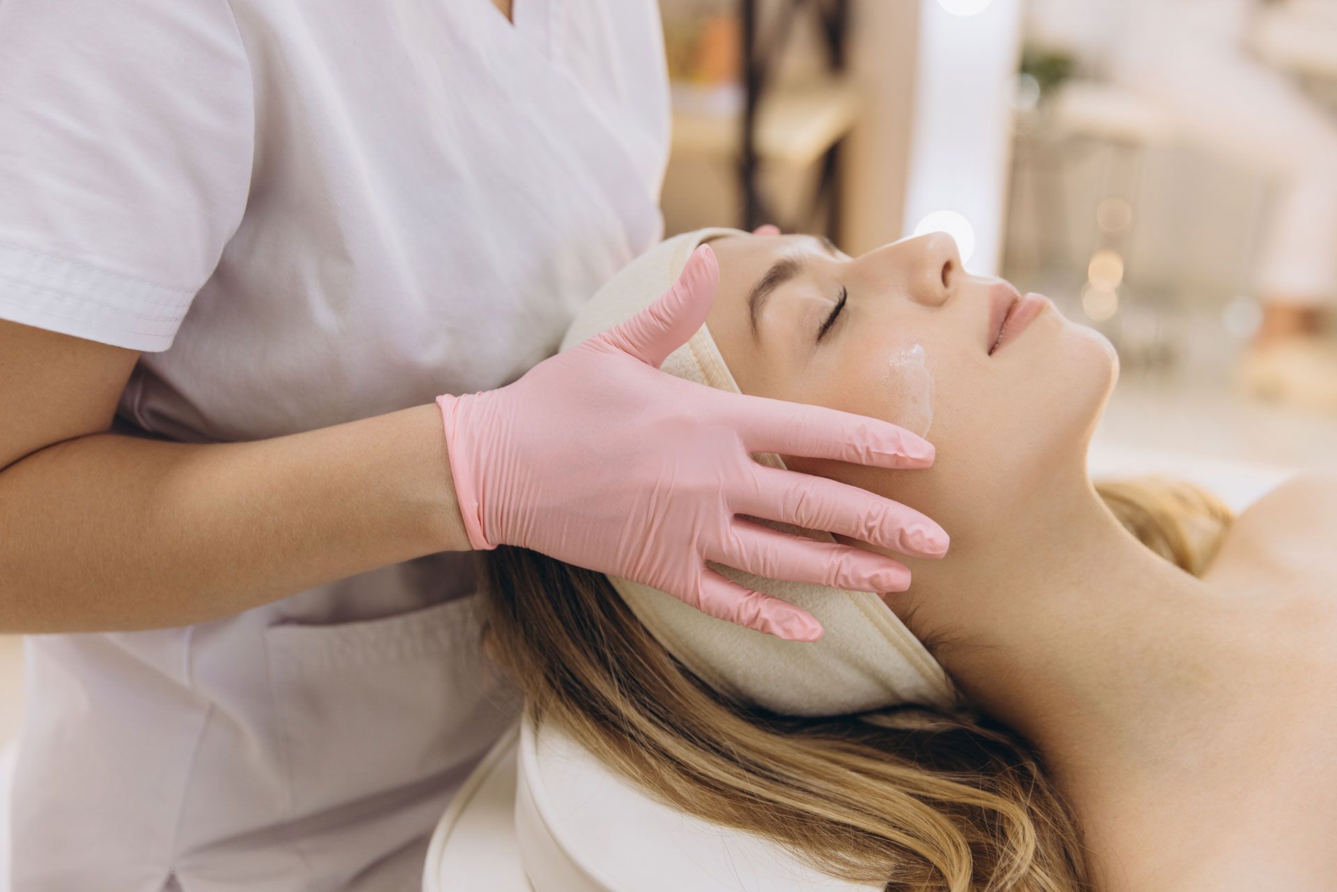 Person receiving facial treatment; pink-gloved hands applying product.