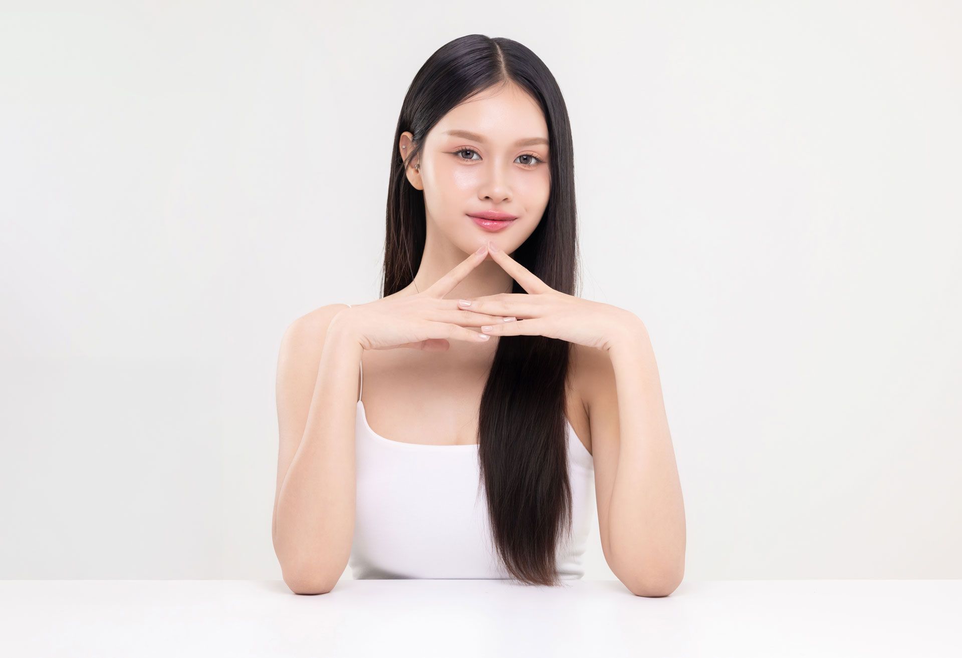 Woman with long black hair, white tank top, hands in front of her face in a triangular shape, white background.