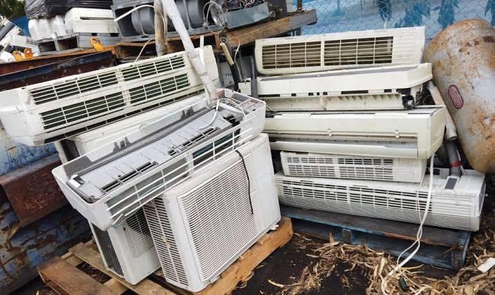 Pile of white air conditioning units in a scrap yard, on a wooden pallet next to a rusty metal container.
