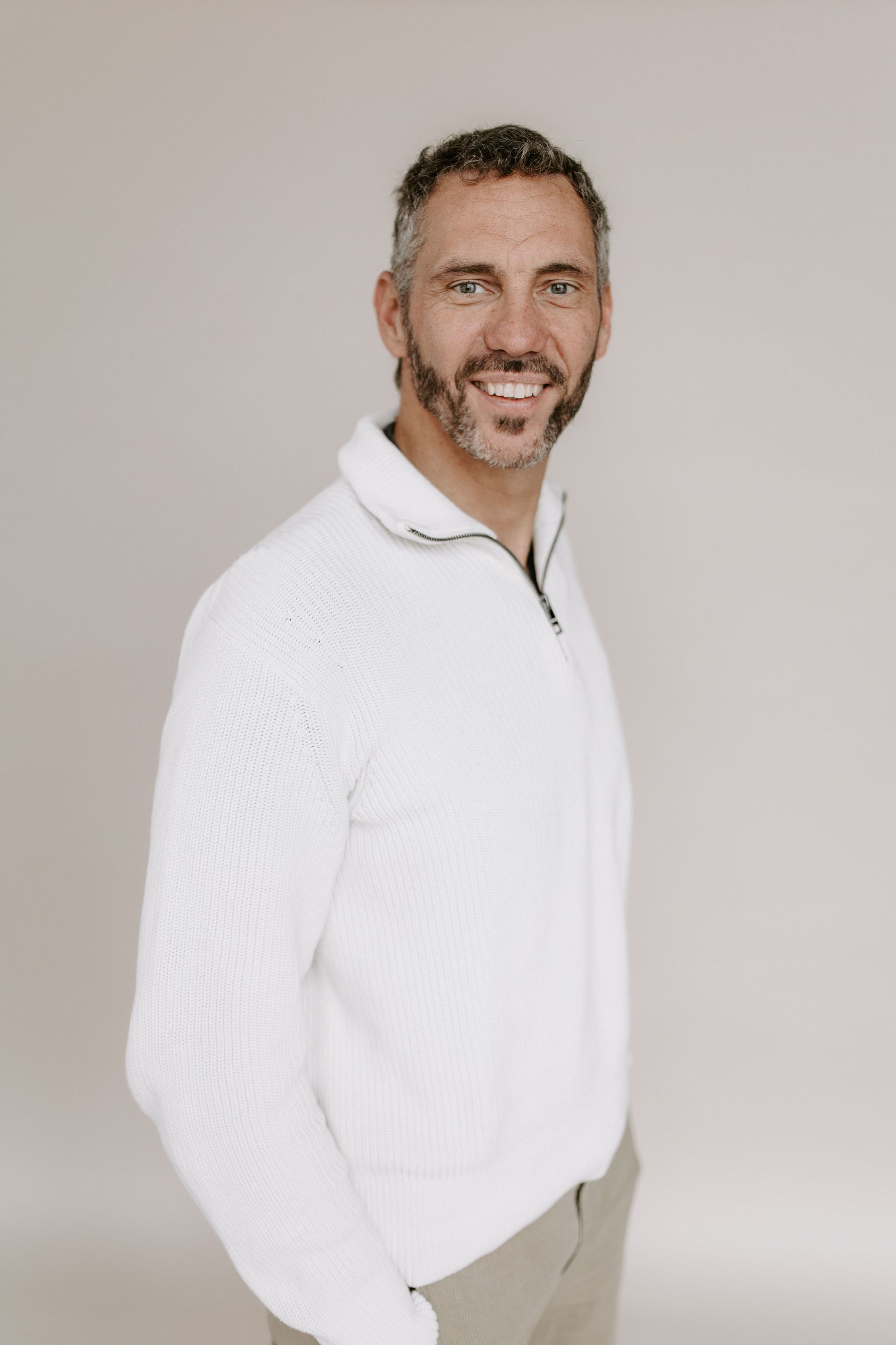 Ein Mann mit Bart trägt einen weißen Pullover und Khakihosen.