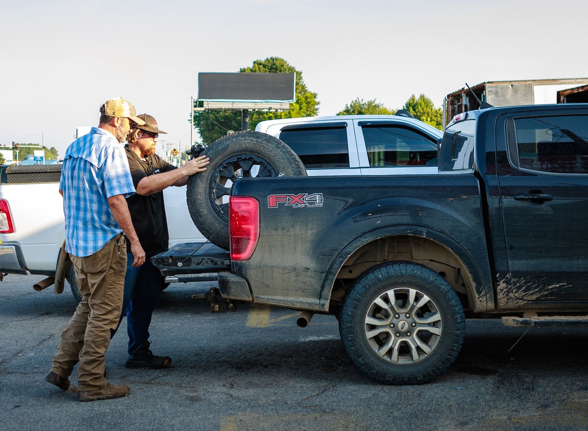 Two people loading a spare tire into the bed of a muddy black pickup truck, outdoors.