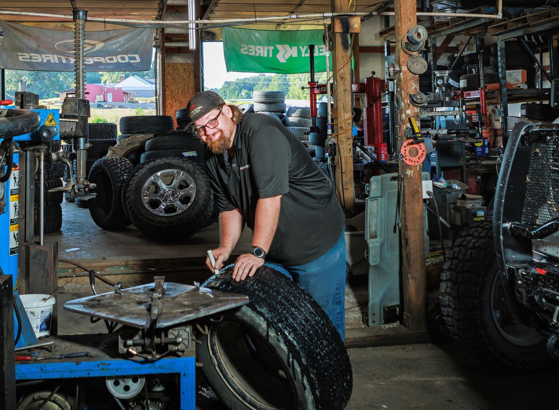 Man in a garage working on a tire, smiling. Tools and tires visible.