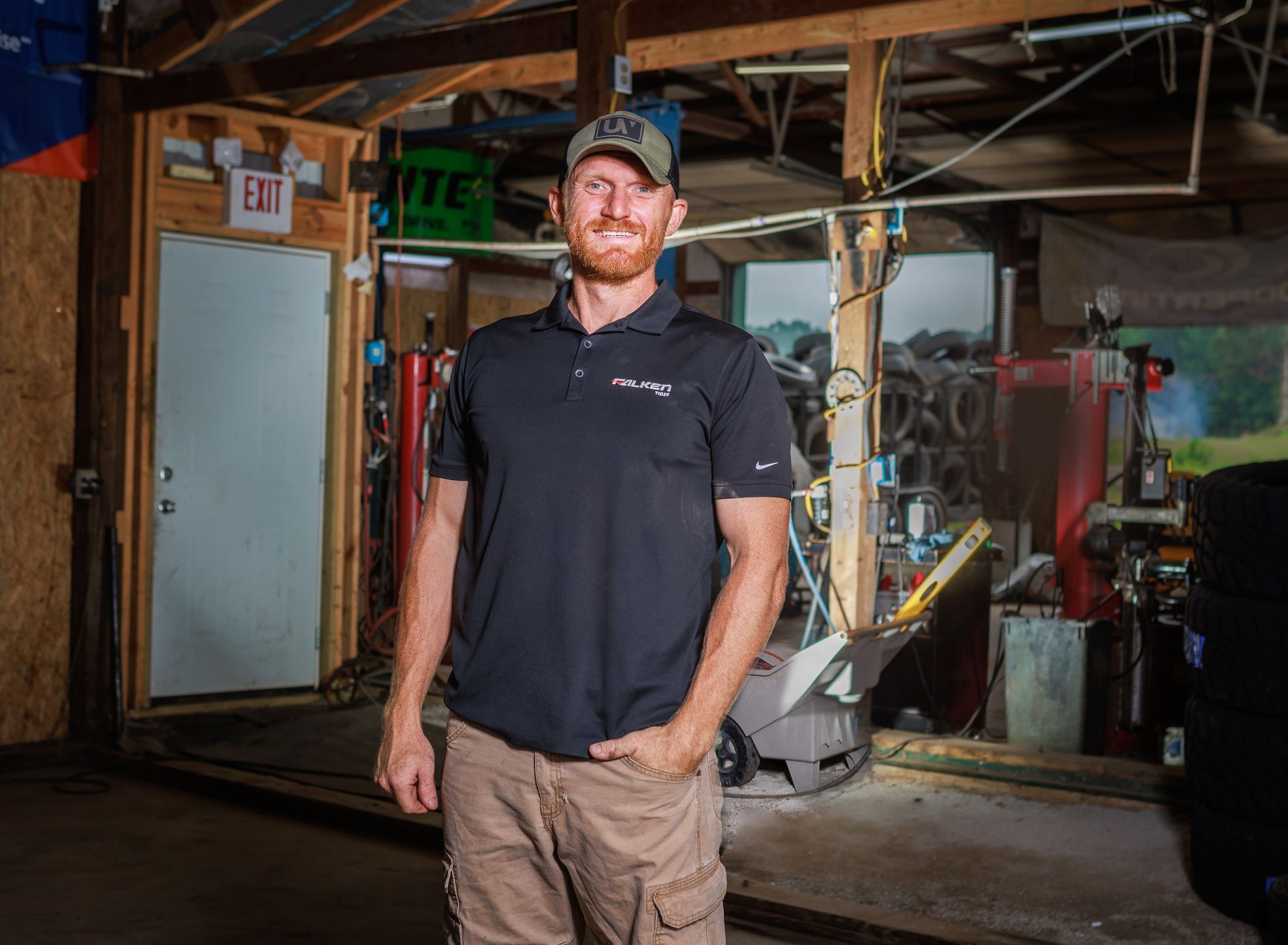 Man in black shirt and cap in a garage; standing with hand in pocket. Tools and tires visible.
