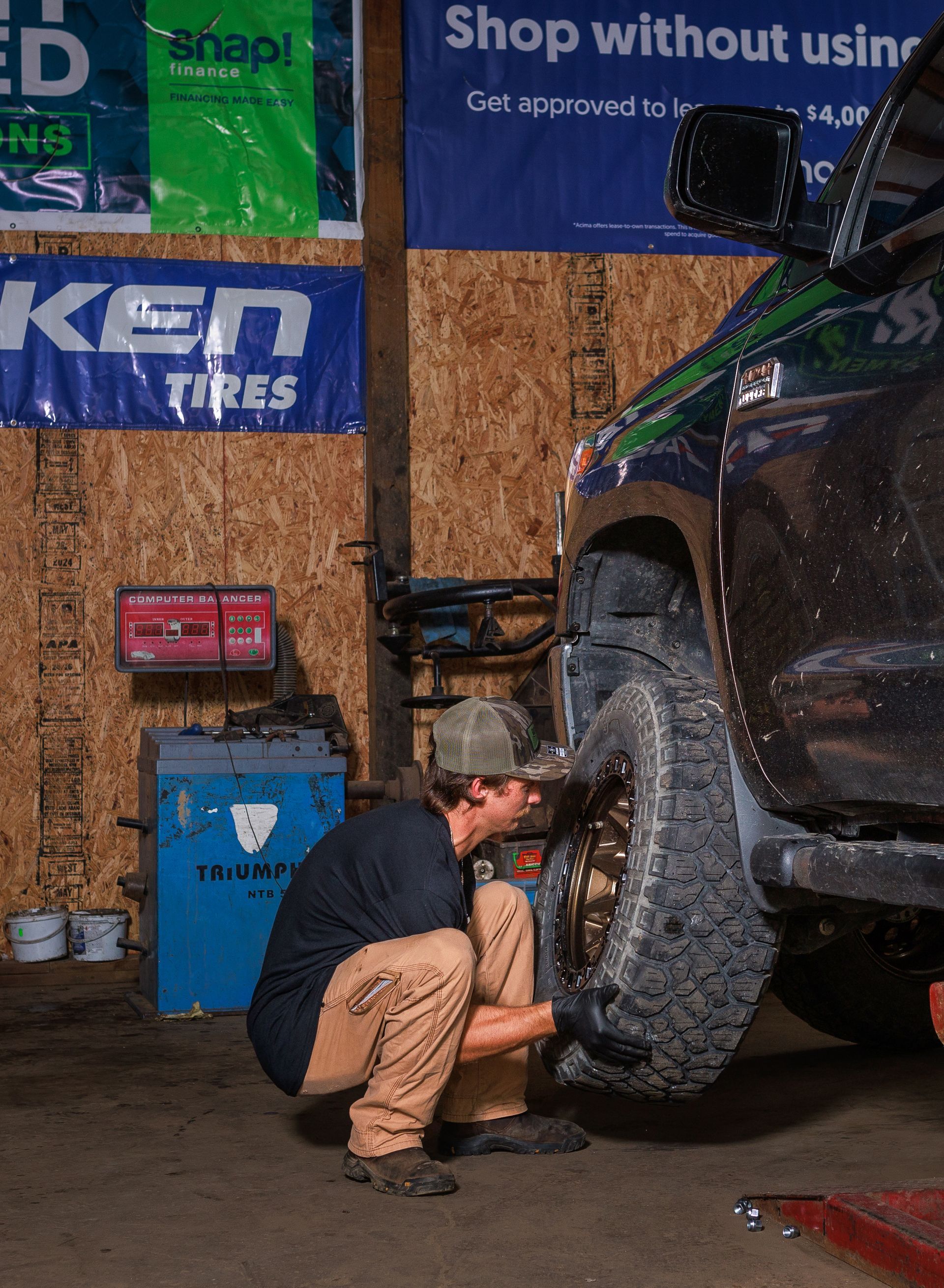 Mechanic changing a tire in a garage. Gold wheel, black truck, brown pants, tools.