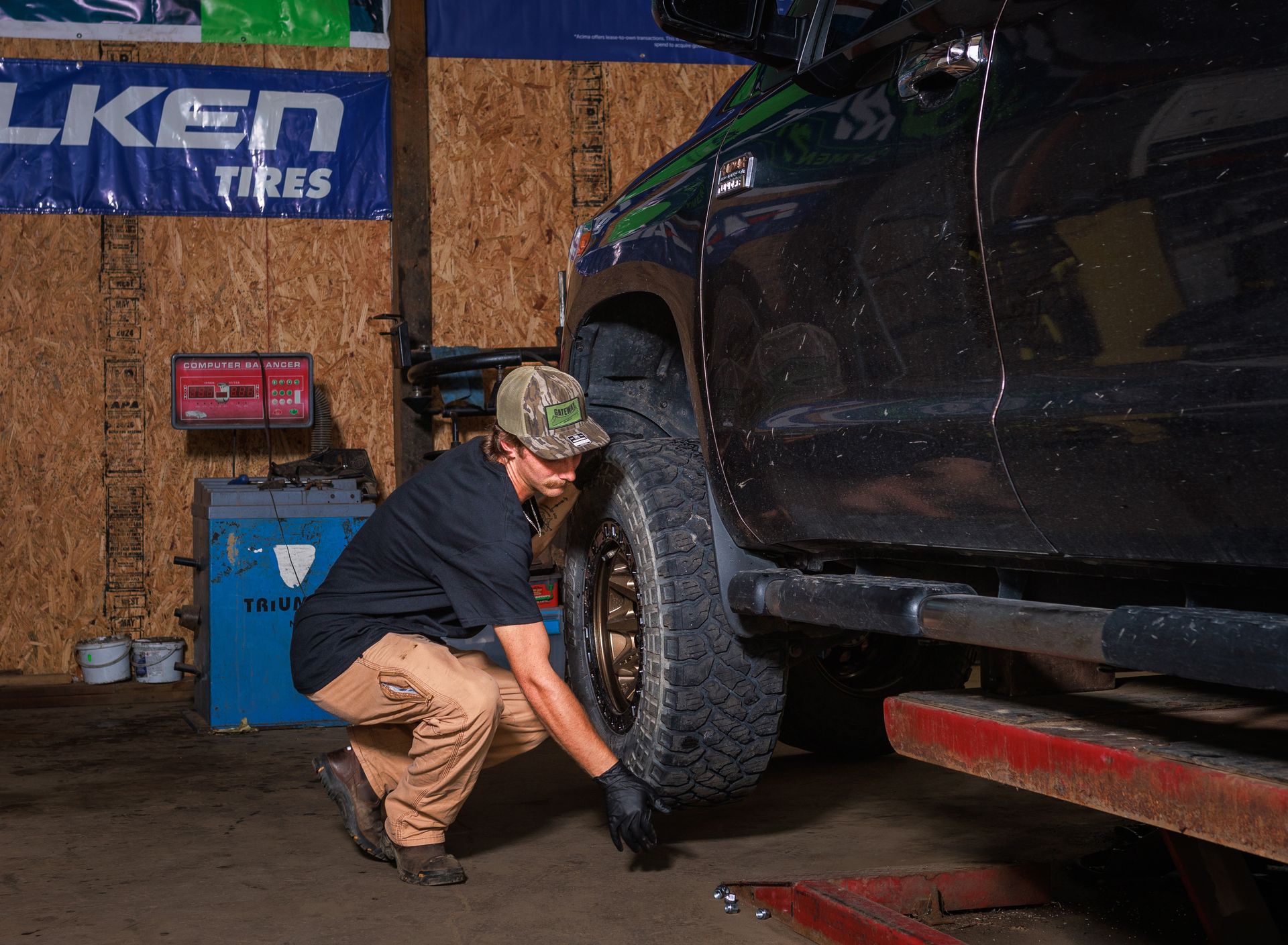 Mechanic inspects tire on a lifted black truck in a garage. Wearing gloves, and a hat.