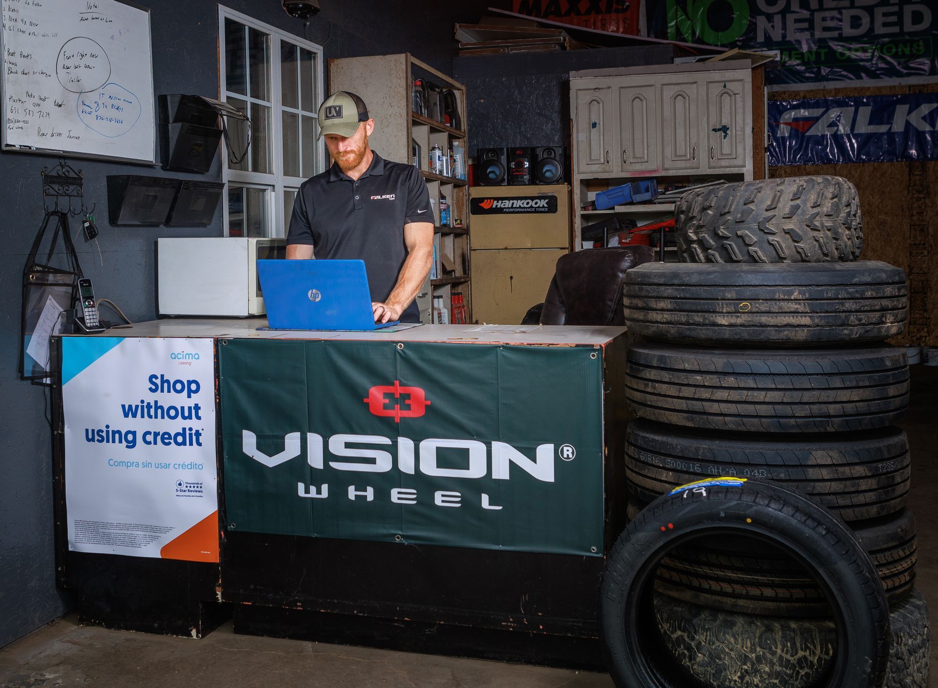 Man working on laptop behind a Vision Wheel counter in a shop; tires stacked nearby.
