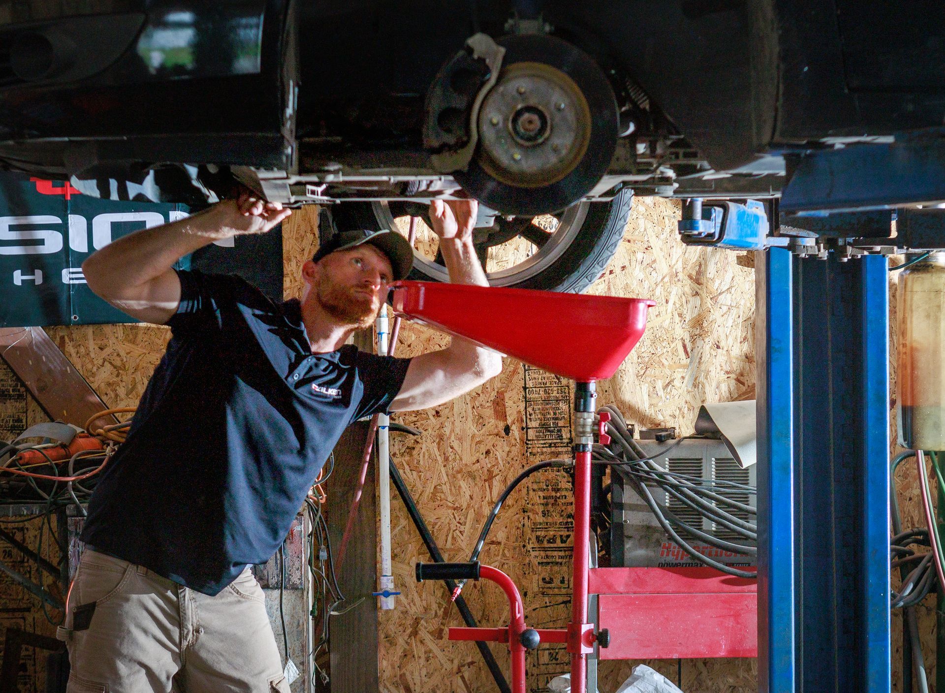 Mechanic under car, draining oil into a red funnel. Car is on a lift in a garage.