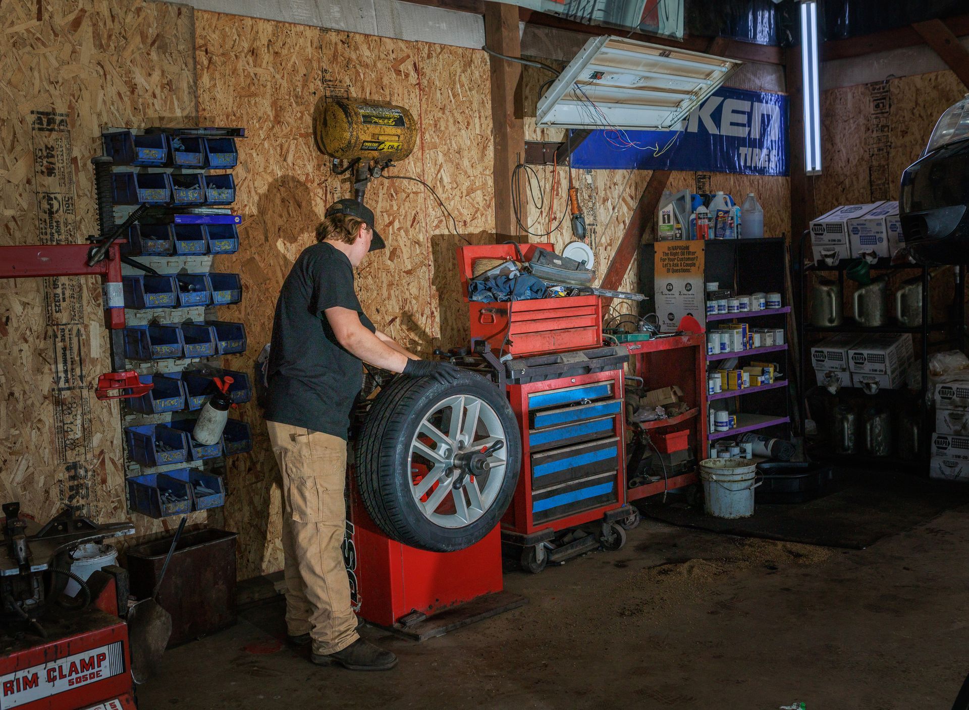 Person balances a tire in a workshop. A red balancing machine and various tools are visible.