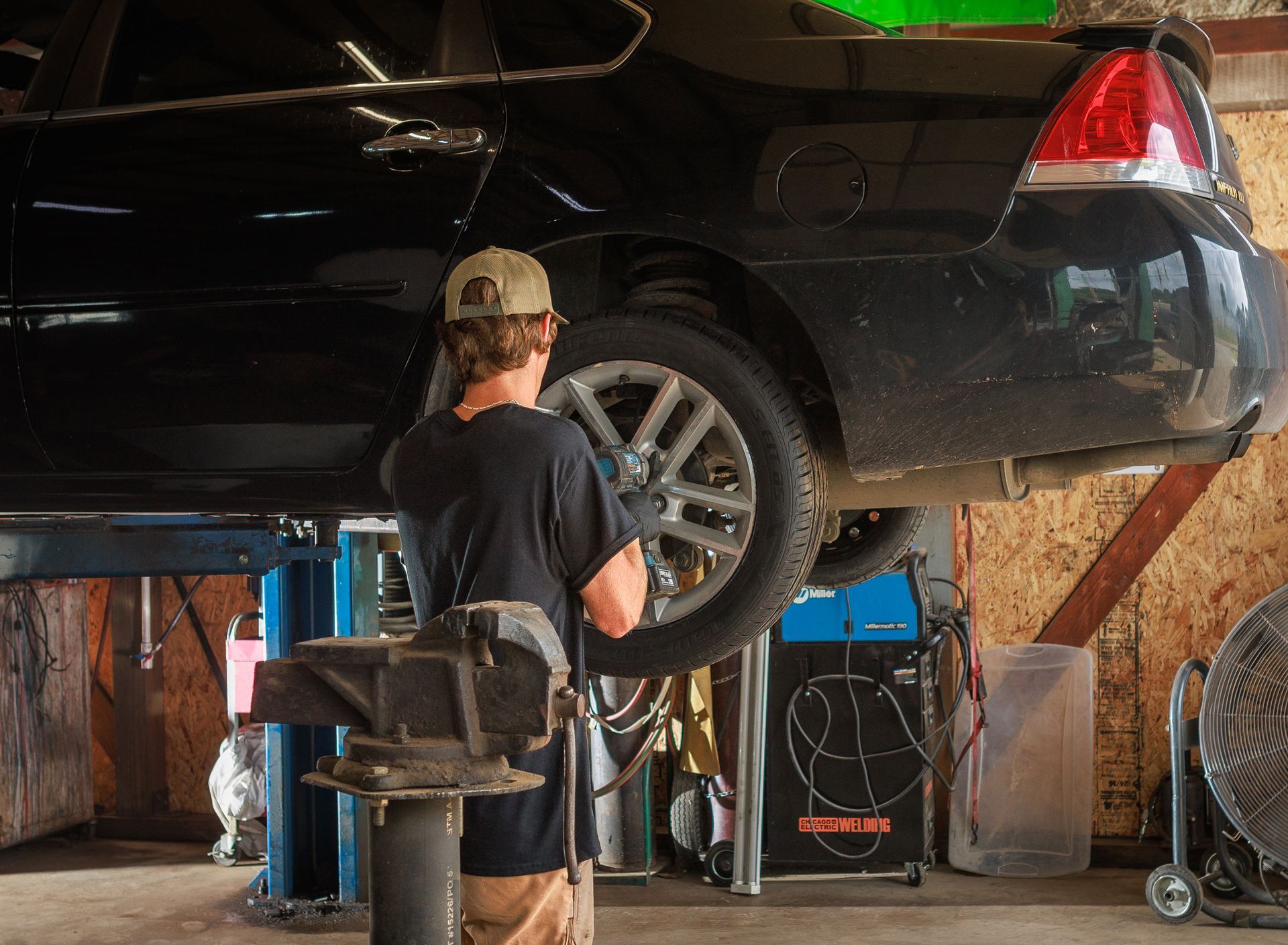 Mechanic working on the raised rear of a black car in a garage, using a tool on the wheel.