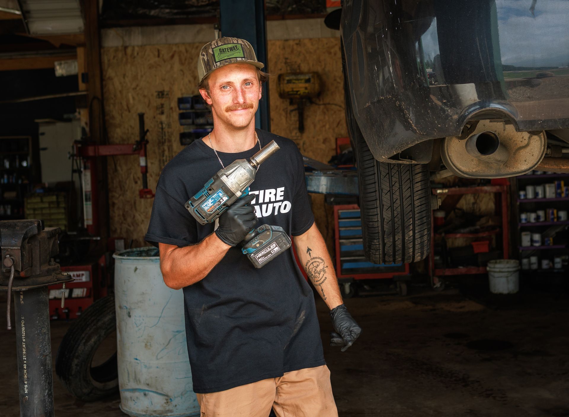 Mechanic holding a power tool, standing in a garage, smiling. Wearing a black shirt, camouflage hat, and gloves.