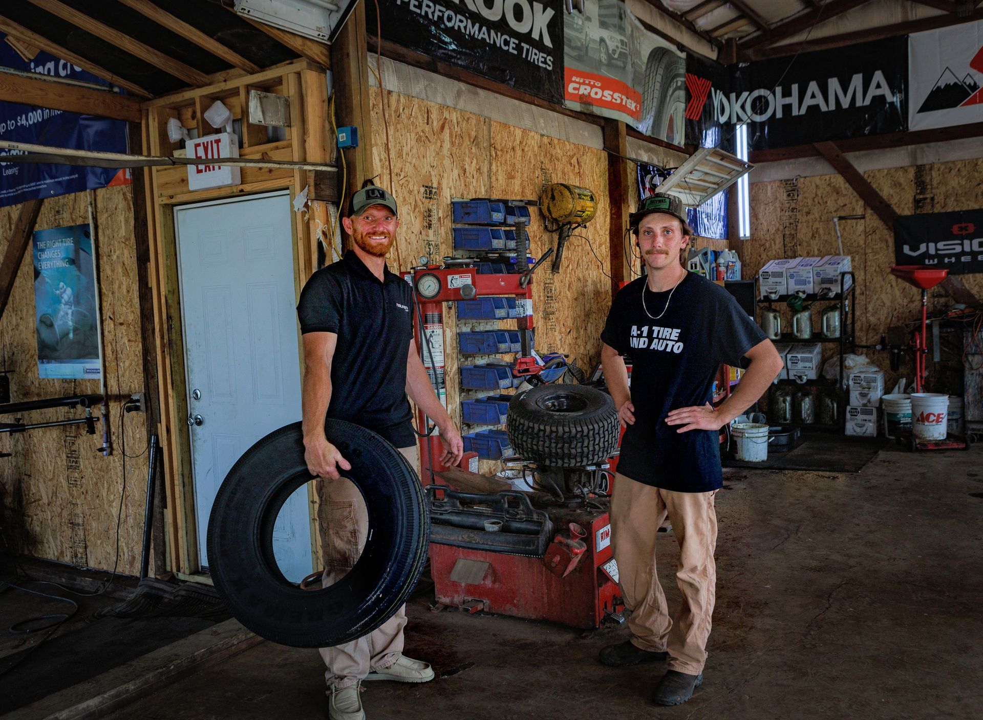Two people in a garage, one holding a tire, near tire changing equipment.