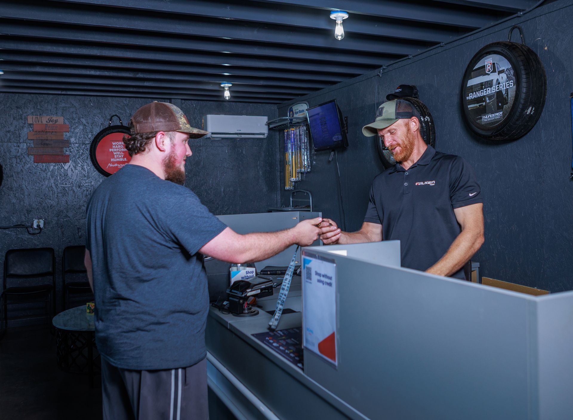 Two men at a counter, one handing the other something. Inside a business with dark walls.