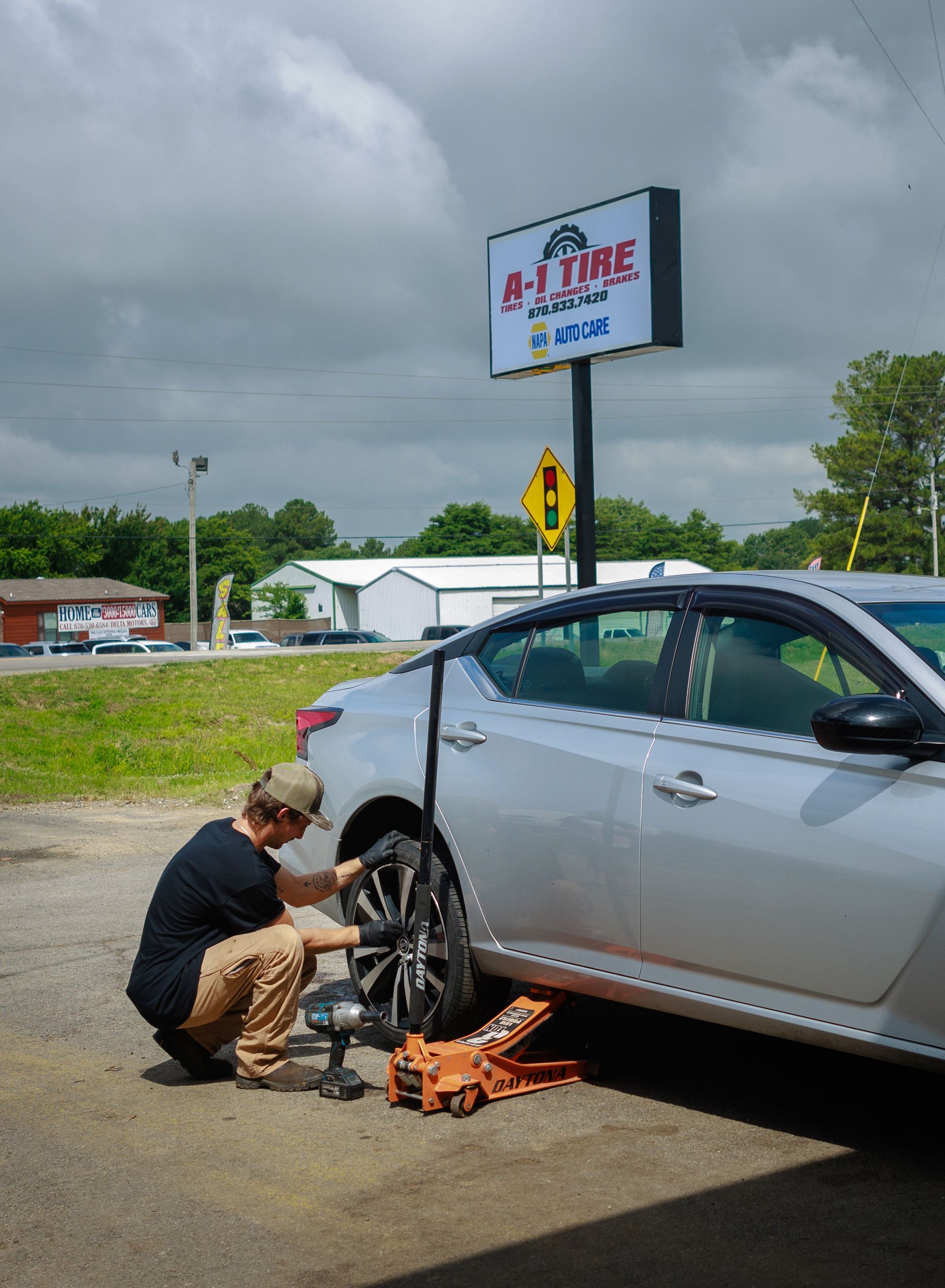 Person changing a tire at a tire shop. Silver car jacked up, wheel removed. Shop sign in background.