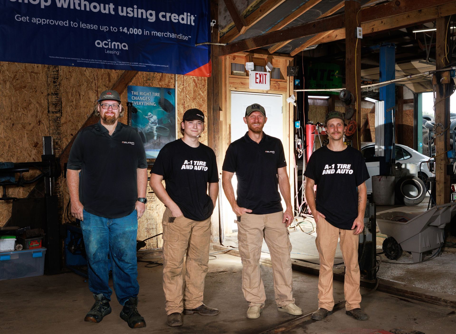 Four people in a garage, wearing black shirts and tan pants. Standing in front of a banner.